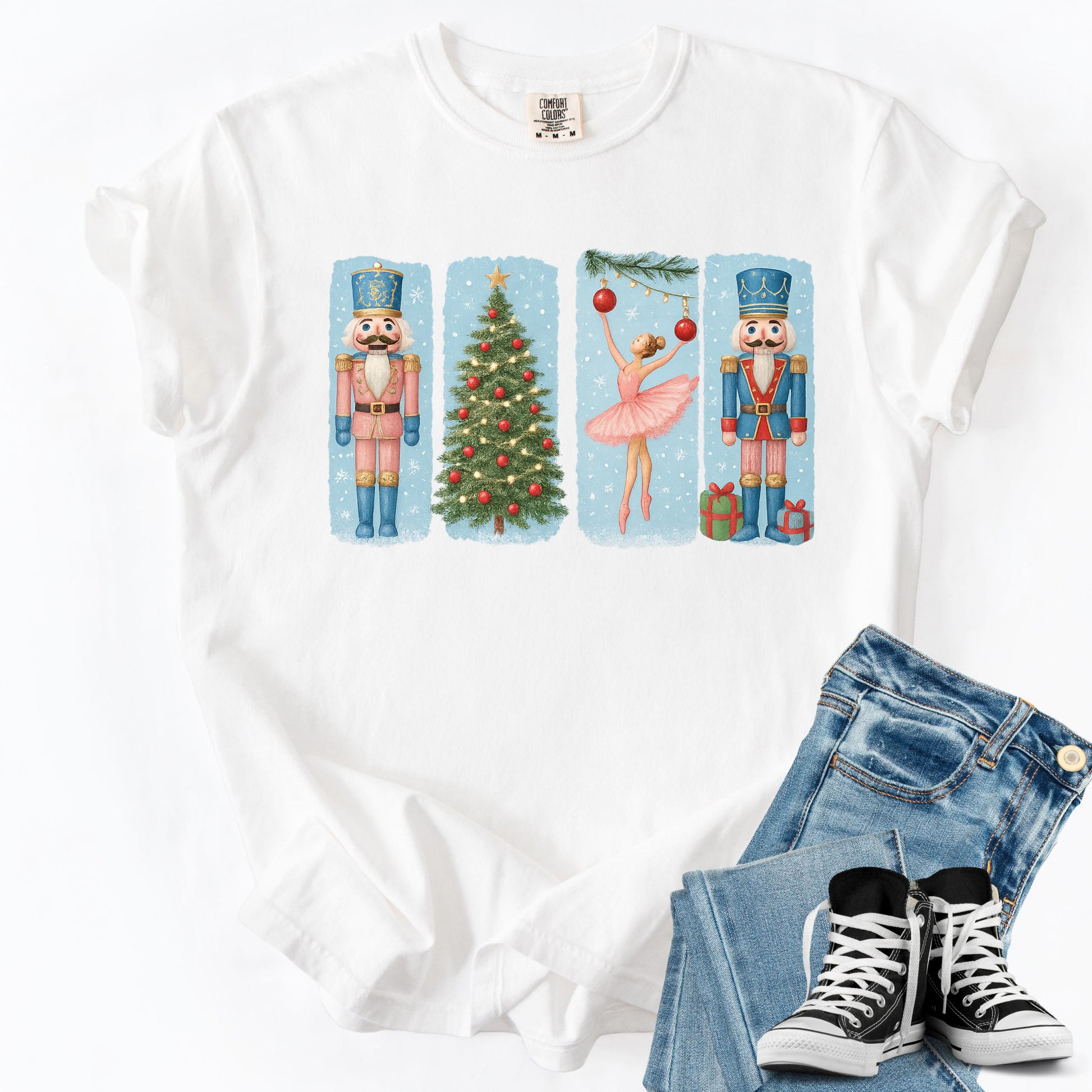 May include: White t-shirt featuring a Christmas-themed design. The design includes a nutcracker soldier in a pink uniform, a decorated Christmas tree, a ballerina in a pink tutu, and another nutcracker soldier in a blue uniform. The design is set against a light blue background.