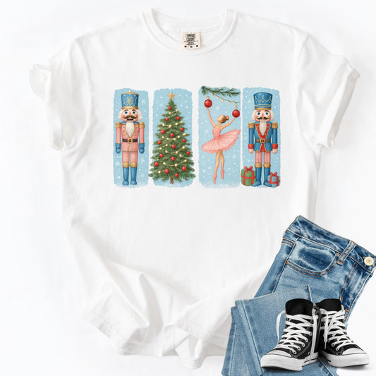 May include: White t-shirt featuring a Christmas-themed design. The design includes a nutcracker soldier in a pink uniform, a decorated Christmas tree, a ballerina in a pink tutu, and another nutcracker soldier in a blue uniform. The design is set against a light blue background.